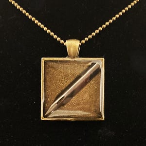 K&ouml;nnte beinhalten: Eine goldene Halskette mit einem quadratischen Anh&auml;nger, der ein goldfarbenes F&uuml;llfederhalter-Spitzen-Design zeigt.