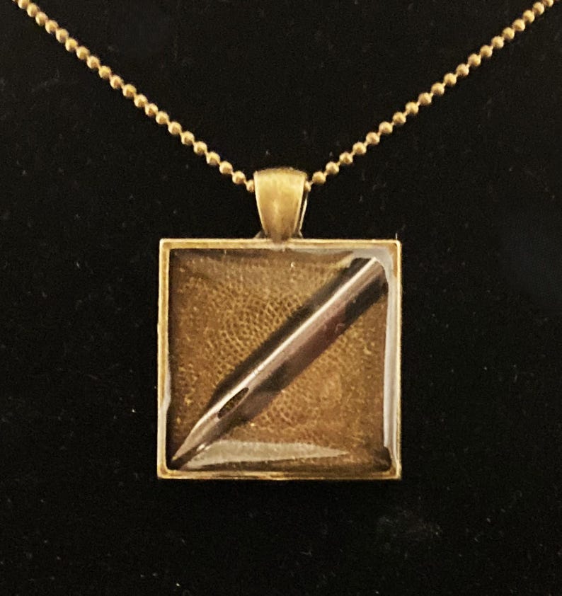 K&ouml;nnte beinhalten: Eine goldene Halskette mit einem quadratischen Anh&auml;nger, der ein goldfarbenes F&uuml;llfederhalter-Spitzen-Design zeigt.
