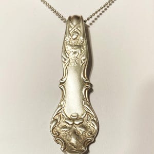 Puede incluir: Un collar de plata con un colgante hecho de una cuchara de plata vintage. La cuchara tiene un dise&ntilde;o ornamentado con motivos florales.