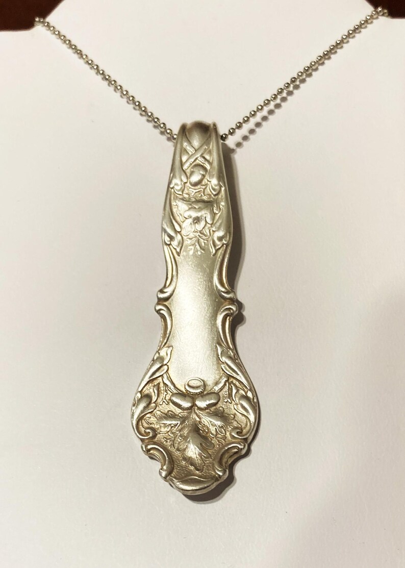 Puede incluir: Un collar de plata con un colgante hecho de una cuchara de plata vintage. La cuchara tiene un dise&ntilde;o ornamentado con motivos florales.