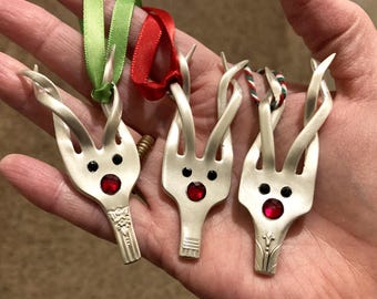 May include: Three silver fork ornaments transformed into reindeer shapes. Each has black eyes and a red gem nose. Green and red ribbons and red and white twine are attached for hanging. A distinctive Christmas decoration.