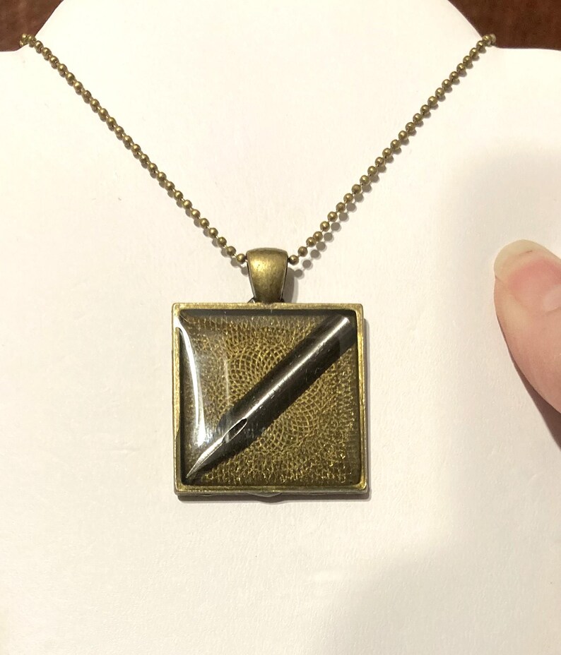 K&ouml;nnte beinhalten: Eine goldene Kettenkette mit einem quadratischen Anh&auml;nger, der eine schwarze Tintenfeder in einem quadratischen, klaren Harz enth&auml;lt.