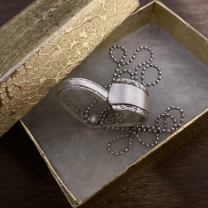 Puede incluir: Un collar con un colgante en forma de coraz&oacute;n de plata con un dise&ntilde;o floral detallado. El collar est&aacute; en una caja de regalo dorada con un patr&oacute;n texturizado.