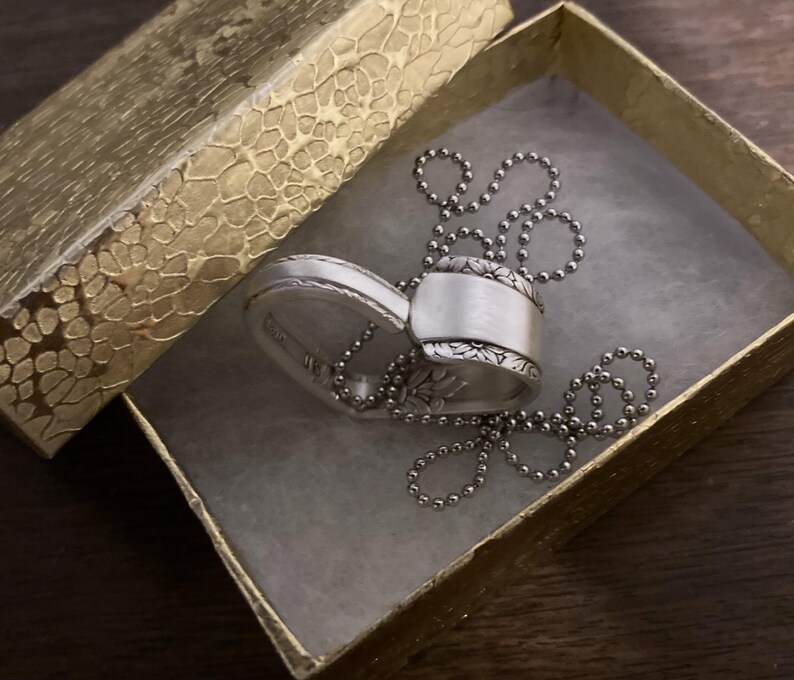 Puede incluir: Un collar con un colgante en forma de coraz&oacute;n de plata con un dise&ntilde;o floral detallado. El collar est&aacute; en una caja de regalo dorada con un patr&oacute;n texturizado.