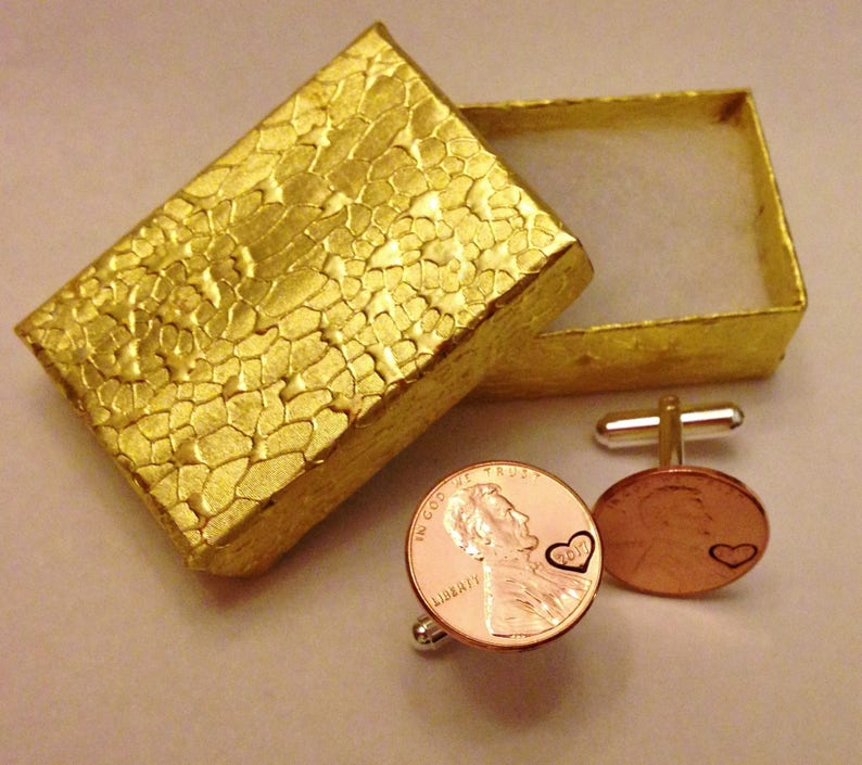 May include: A pair of cufflinks made from a copper penny with a heart design. The cufflinks are in a small gold box with a textured surface.
