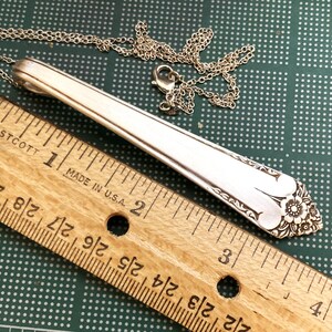 May include: Silver pendant necklace made from a spoon. The pendant is approximately 3.5 inches long and features a floral design at the end. The necklace chain is silver and has a lobster clasp. A wooden ruler is in the foreground.
