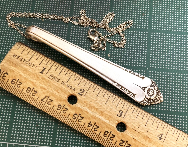 May include: Silver pendant necklace made from a spoon. The pendant is approximately 3.5 inches long and features a floral design at the end. The necklace chain is silver and has a lobster clasp. A wooden ruler is in the foreground.