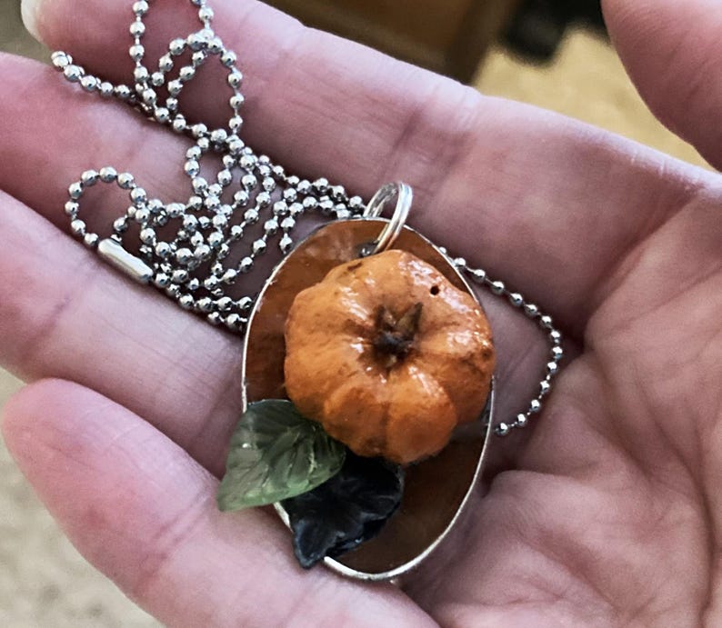 May include: A silver chain necklace with a pendant featuring a small orange pumpkin with green leaves. The pumpkin is nestled in a silver oval-shaped frame.