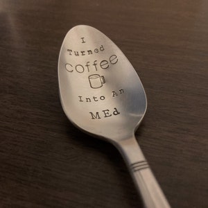 Pu&ograve; includere: Un cucchiaio in acciaio inossidabile con la scritta stampata "I turned Coffee Into An MED" e una grafica di tazza di caff&egrave;. Il cucchiaio &egrave; su una superficie marrone scuro.