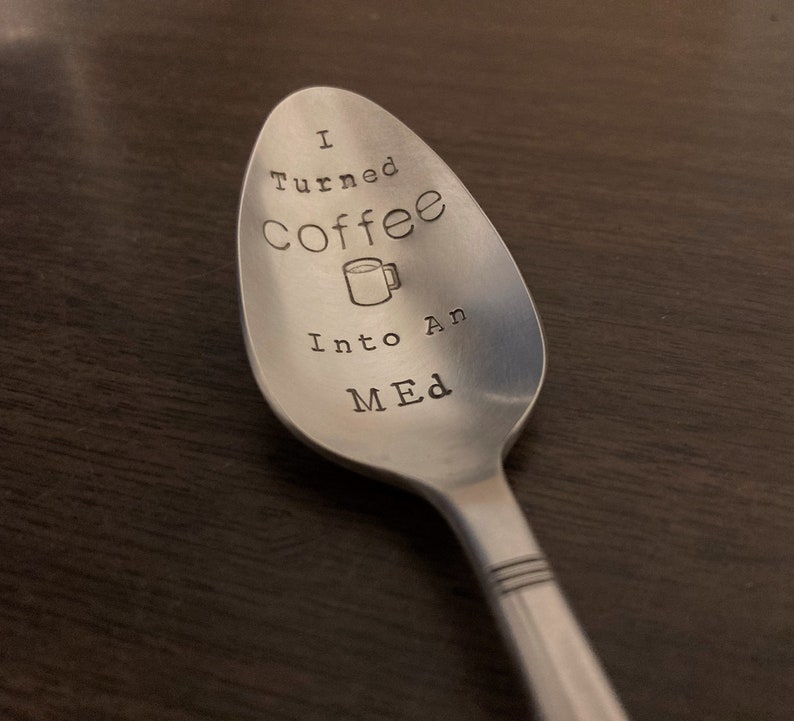Pu&ograve; includere: Un cucchiaio in acciaio inossidabile con la scritta stampata "I turned Coffee Into An MED" e una grafica di tazza di caff&egrave;. Il cucchiaio &egrave; su una superficie marrone scuro.