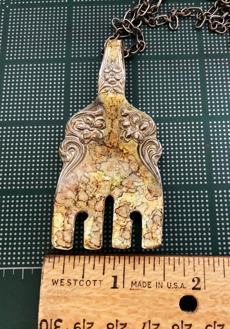 May include: A silver fork necklace with a decorative handle and tarnished finish. The fork is about 1 inch long. The necklace is made of a silver chain.