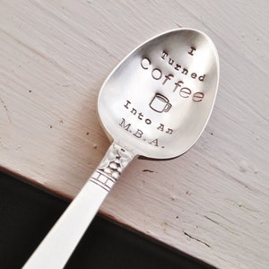 Pu&ograve; includere: Un cucchiaio d'argento con la scritta stampata "I turned coffee into an M.B.A." e una piccola grafica di tazza di caff&egrave;. Il cucchiaio ha un manico decorativo ed &egrave; appoggiato su una superficie di legno chiaro.