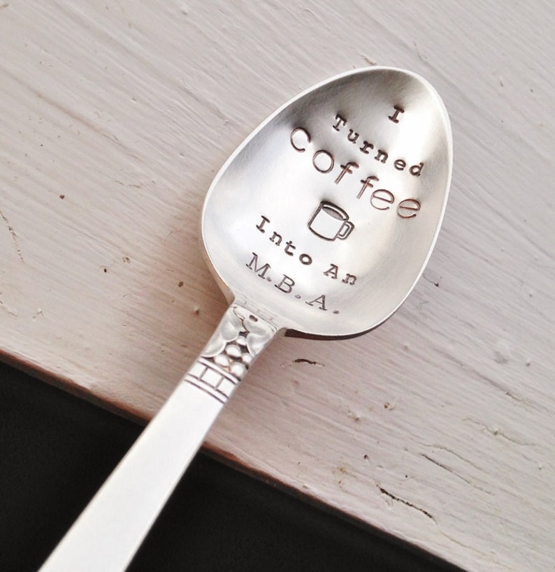Pu&ograve; includere: Un cucchiaio d'argento con la scritta stampata "I turned coffee into an M.B.A." e una piccola grafica di tazza di caff&egrave;. Il cucchiaio ha un manico decorativo ed &egrave; appoggiato su una superficie di legno chiaro.