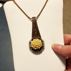May include: A necklace with a copper chain and a pendant made from a vintage spoon with a cream-coloured flower charm.
