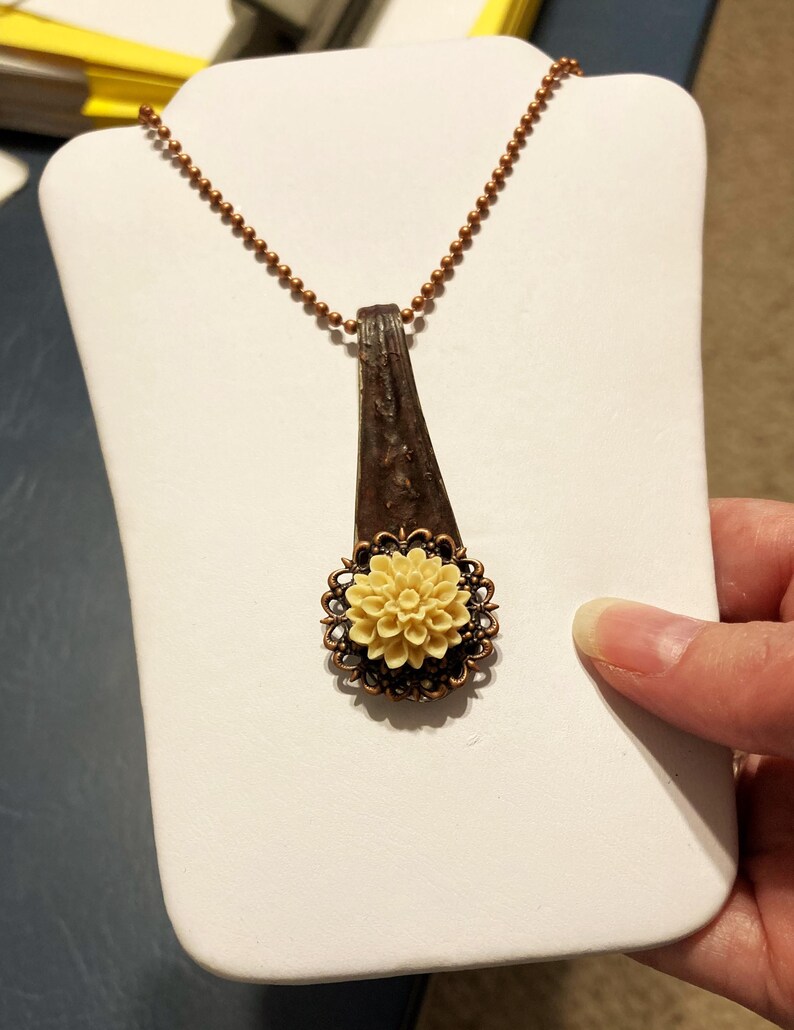 May include: A necklace with a copper chain and a pendant made from a vintage spoon with a cream-coloured flower charm.