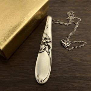 Puede incluir: Un collar con colgante plateado hecho de una cuchara, con detalles florales. El colgante est&aacute; conectado a una delicada cadena. Una caja dorada es parcialmente visible en el fondo, sobre una superficie de madera oscura.