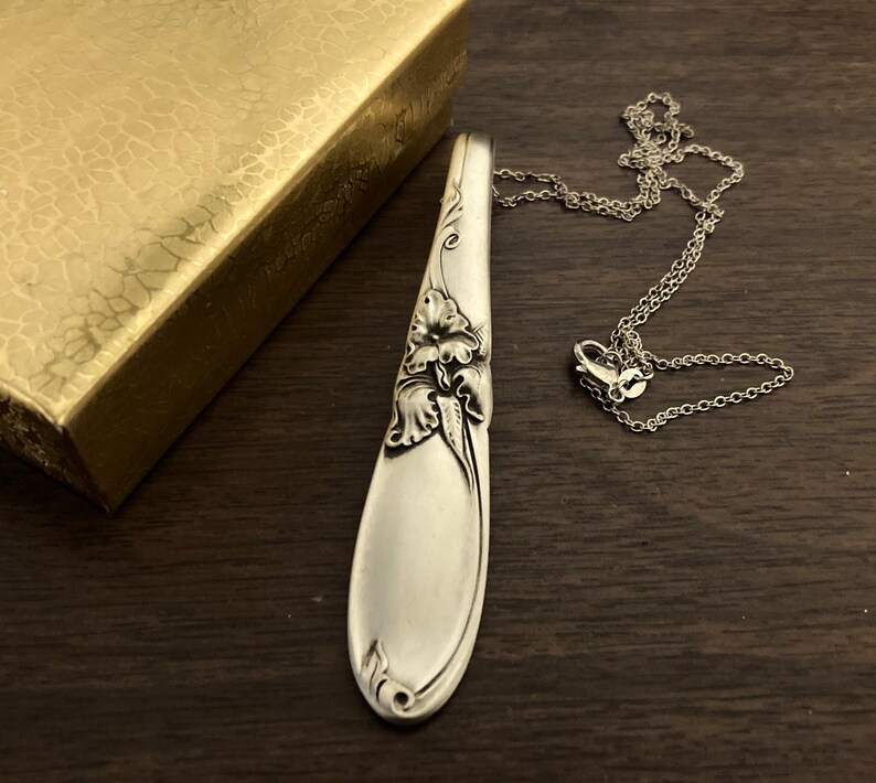 Puede incluir: Un collar con colgante plateado hecho de una cuchara, con detalles florales. El colgante est&aacute; conectado a una delicada cadena. Una caja dorada es parcialmente visible en el fondo, sobre una superficie de madera oscura.