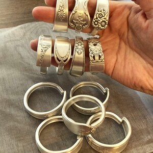 Puede incluir: Una colecci&oacute;n de anillos de cuchara de plata, algunos con dise&ntilde;os florales, otros con bandas lisas. Los anillos est&aacute;n dispuestos en dos filas, con la fila superior mostrando los anillos en una mano y la fila inferior mostrando los anillos apilados en una mesa.