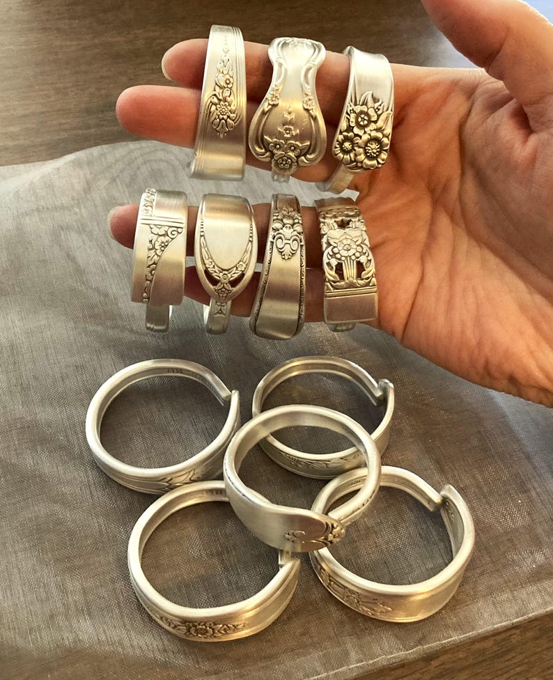 Puede incluir: Una colecci&oacute;n de anillos de cuchara de plata, algunos con dise&ntilde;os florales, otros con bandas lisas. Los anillos est&aacute;n dispuestos en dos filas, con la fila superior mostrando los anillos en una mano y la fila inferior mostrando los anillos apilados en una mesa.