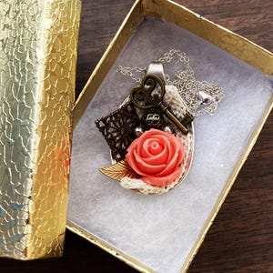 May include: A silver spoon necklace with a coral rose, a key, and a lace design. The necklace is in a small gift box.