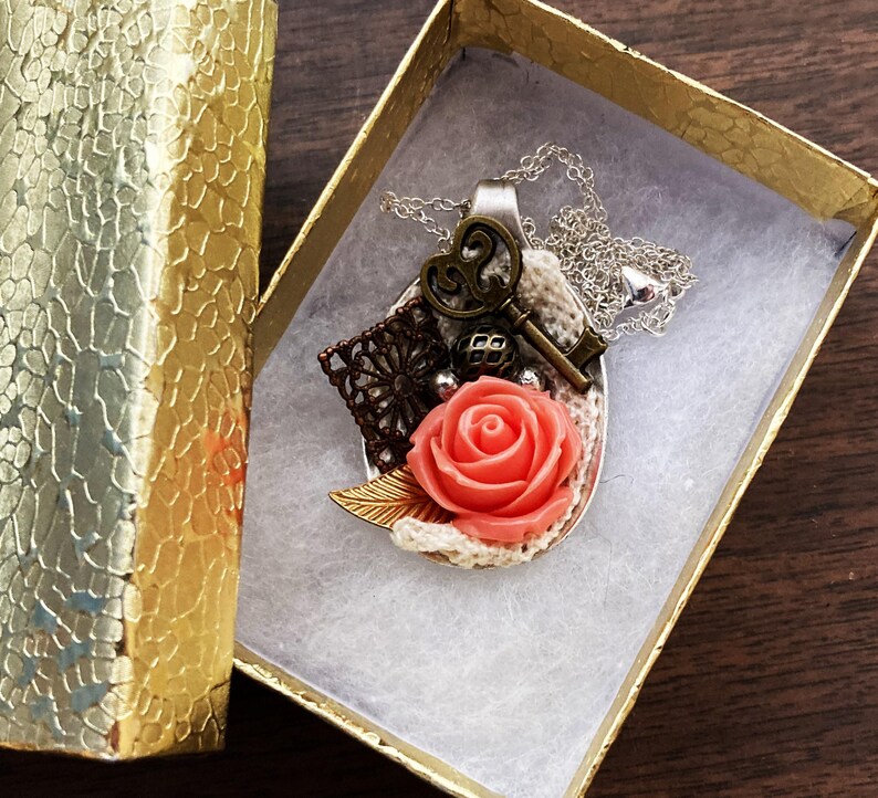 May include: A silver spoon necklace with a coral rose, a key, and a lace design. The necklace is in a small gift box.