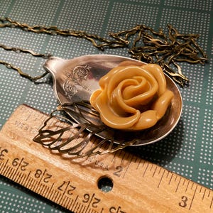 May include: A gold-tone necklace with a vintage spoon pendant featuring a brown resin rose. The spoon has an ornate design and is attached to a delicate chain.