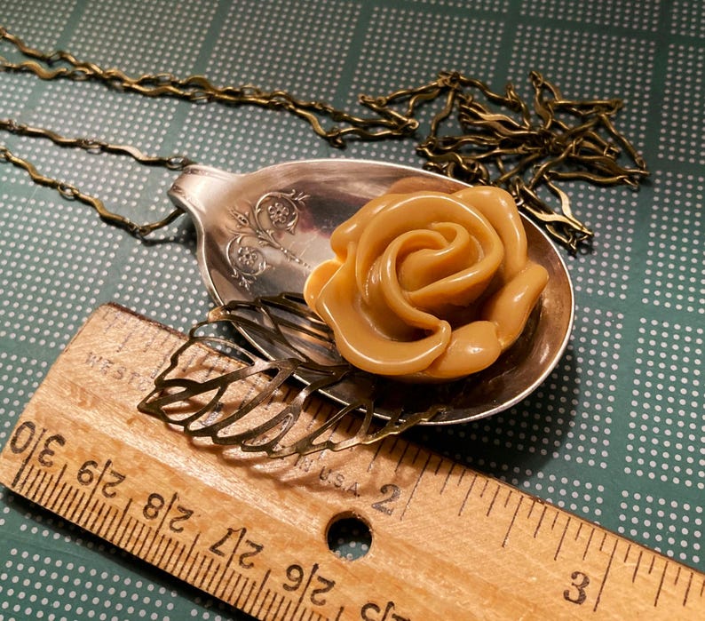 May include: A gold-tone necklace with a vintage spoon pendant featuring a brown resin rose. The spoon has an ornate design and is attached to a delicate chain.