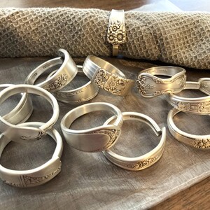 Puede incluir: Una colecci&oacute;n de anillos de servilleta plateados hechos con cubiertos reutilizados. Los anillos presentan intrincados dise&ntilde;os florales y est&aacute;n dispuestos sobre una superficie de tela marr&oacute;n claro.