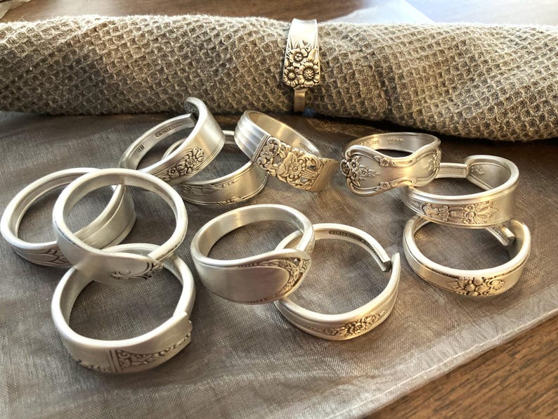Puede incluir: Una colecci&oacute;n de anillos de servilleta plateados hechos con cubiertos reutilizados. Los anillos presentan intrincados dise&ntilde;os florales y est&aacute;n dispuestos sobre una superficie de tela marr&oacute;n claro.