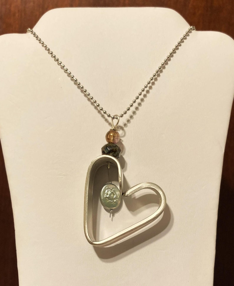 May include: A silver heart-shaped pendant necklace with a silver chain. The pendant has a smaller circle inside of it with a green and white design. The necklace also has three beads in brown, pink, and green.