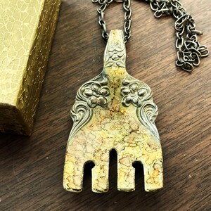 May include: A silver fork with ornate floral designs has been repurposed into a necklace. The fork has a yellow patina and is hanging from a silver chain.