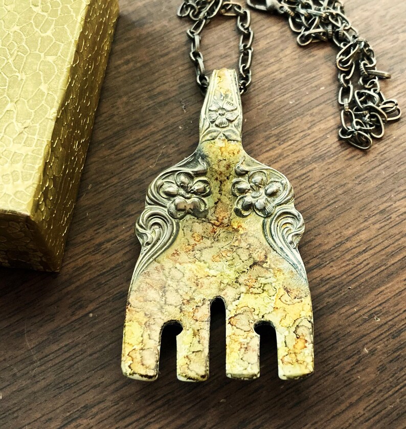 May include: A silver fork with ornate floral designs has been repurposed into a necklace. The fork has a yellow patina and is hanging from a silver chain.