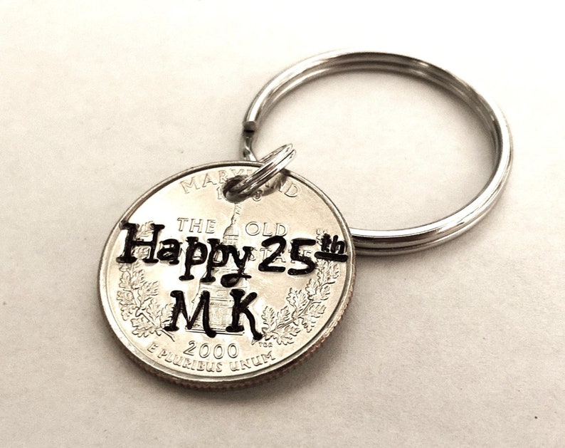 May include: A silver quarter coin keychain with the text "Happy 25th MK" stamped on it. The keychain includes a silver split ring. The coin is from the year 2000 and features the state of Maryland.