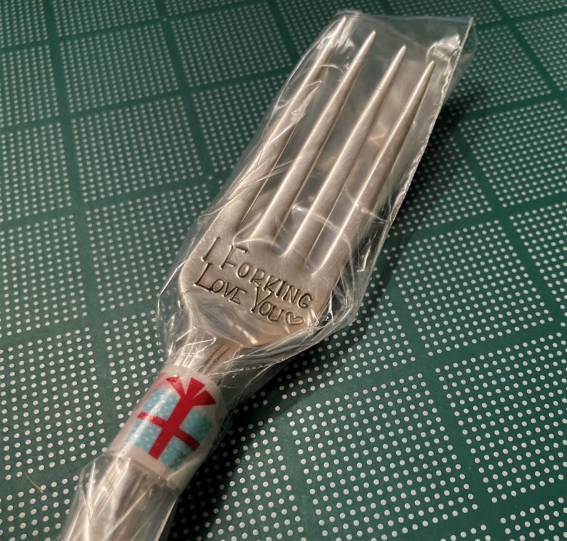 Op de afbeelding: Een zilveren vork met de woorden "I Forking Love You" gegraveerd op het handvat. De vork is verpakt in doorzichtig plastic en vastgebonden met een blauw en rood cadeau-etiket.