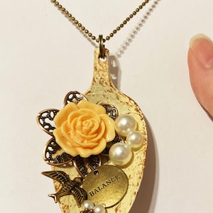 May include: A white spoon necklace with a peach-colored flower, pearl accents, and a gold charm that says "BALANCE".