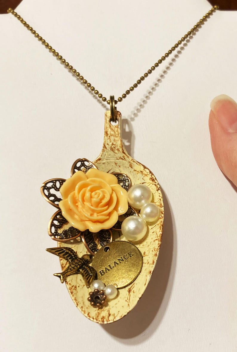 May include: A white spoon necklace with a peach-colored flower, pearl accents, and a gold charm that says "BALANCE".