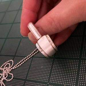 Puede incluir: Un collar con un colgante de cuchara de plata con un dise&ntilde;o adornado. El colgante est&aacute; unido a una cadena de plata.