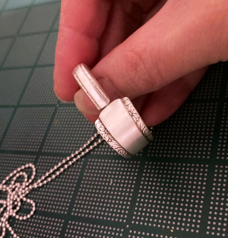Puede incluir: Un collar con un colgante de cuchara de plata con un dise&ntilde;o adornado. El colgante est&aacute; unido a una cadena de plata.