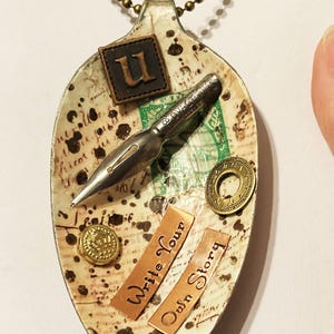 Op de afbeelding: Een witte lepelketting met een vintage pen, een 'U'-bedel en de woorden 'Schrijf je eigen verhaal'.