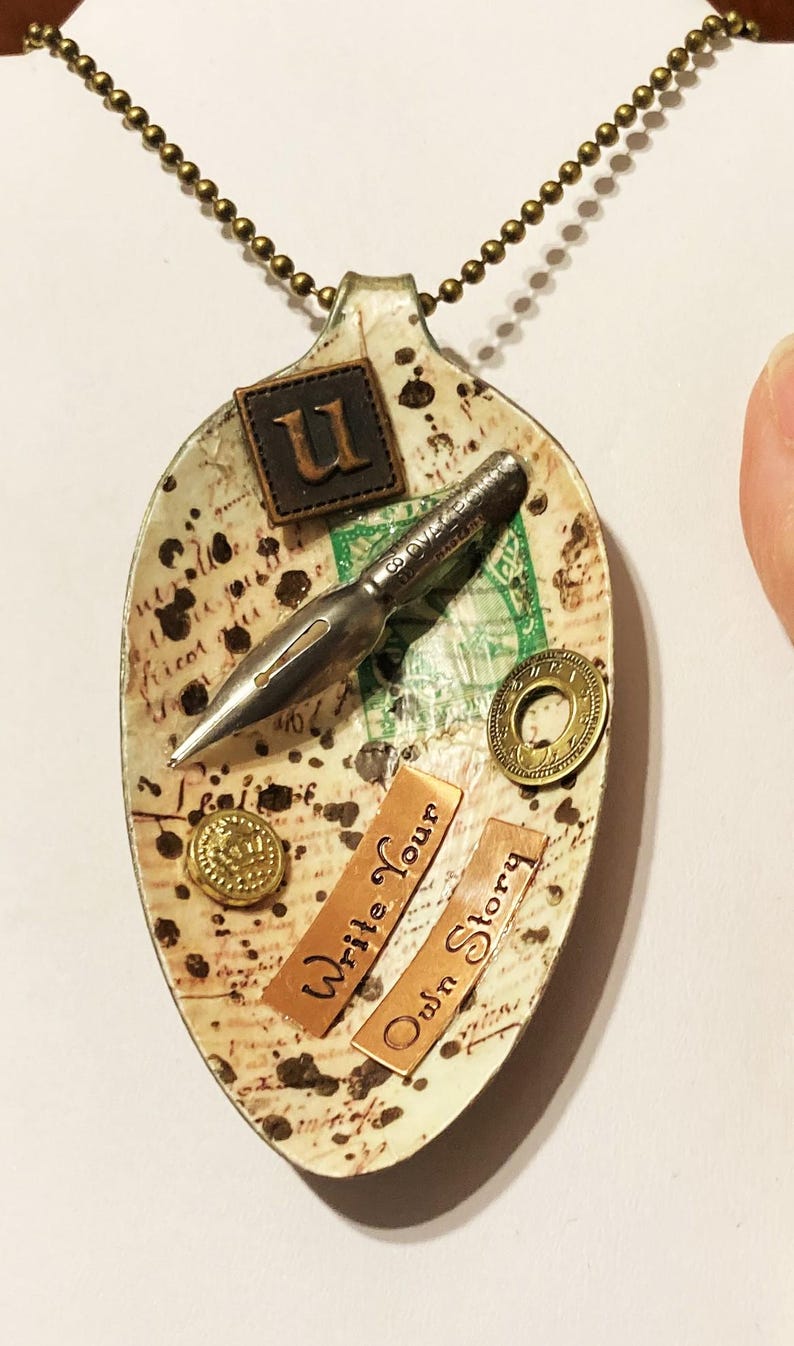 Op de afbeelding: Een witte lepelketting met een vintage pen, een 'U'-bedel en de woorden 'Schrijf je eigen verhaal'.