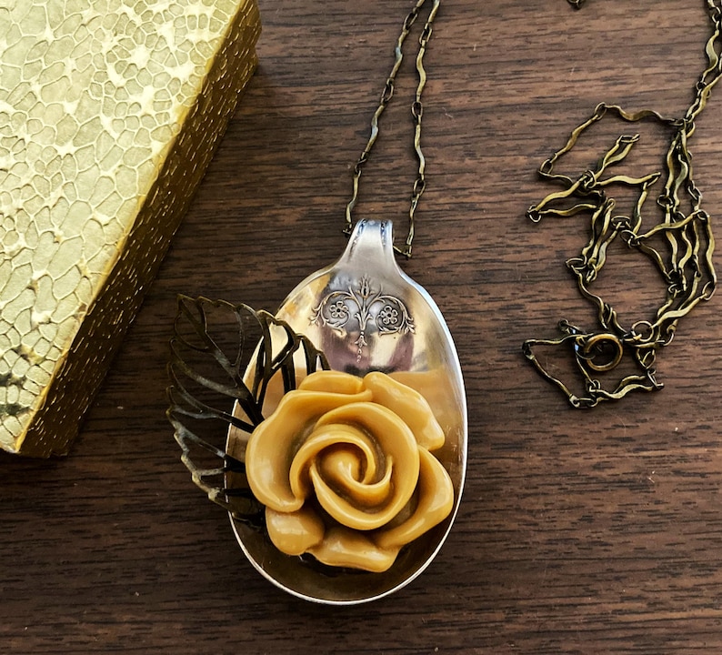 May include: A gold-toned rose made of polymer clay is nestled in a vintage silver spoon pendant. The spoon is attached to a gold chain necklace. The pendant is surrounded by a delicate, openwork leaf design.