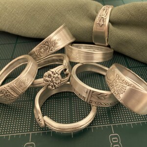 May include: A set of six silver napkin rings made from repurposed silverware. Each ring features a different ornate design.