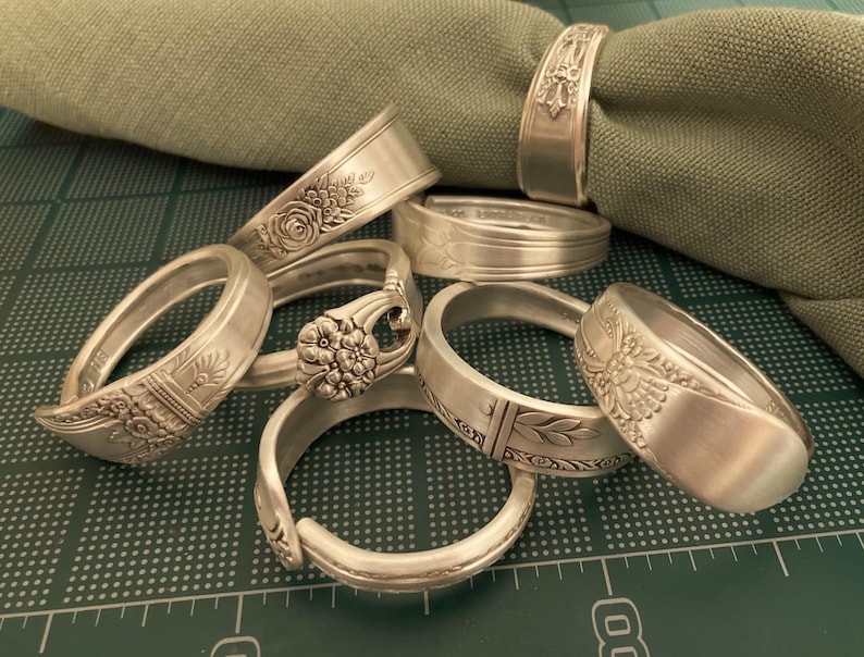 May include: A set of six silver napkin rings made from repurposed silverware. Each ring features a different ornate design.