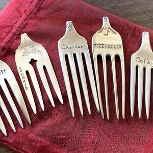 May include: Six silver forks with different cheese names engraved on them. The forks are arranged on a red fabric surface.