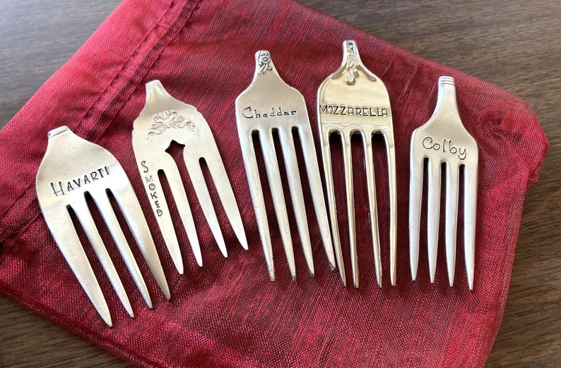 May include: Six silver forks with different cheese names engraved on them. The forks are arranged on a red fabric surface.