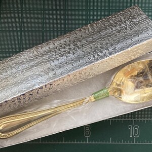 May include: A gold-colored spoon with a green band on the handle, presented in a silver patterned box. The spoon is inside a white box, and the handle has a decorative design. The box is rectangular.