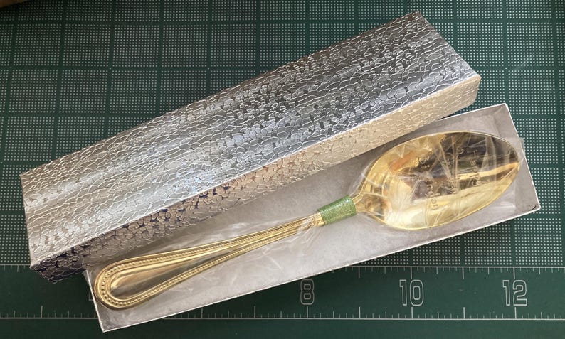 May include: A gold-colored spoon with a green band on the handle, presented in a silver patterned box. The spoon is inside a white box, and the handle has a decorative design. The box is rectangular.