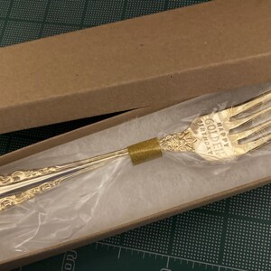 May include: A gold-colored fork with ornate detailing, presented in a cardboard box. The fork has the words "HAPPY GOLDEN BIRTHDAY" engraved on it. The fork is wrapped in clear plastic and rests on a white surface.