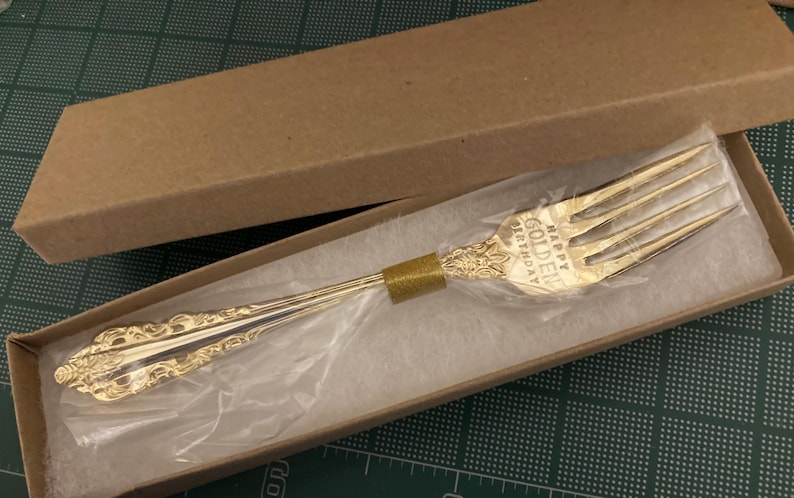 May include: A gold-colored fork with ornate detailing, presented in a cardboard box. The fork has the words "HAPPY GOLDEN BIRTHDAY" engraved on it. The fork is wrapped in clear plastic and rests on a white surface.
