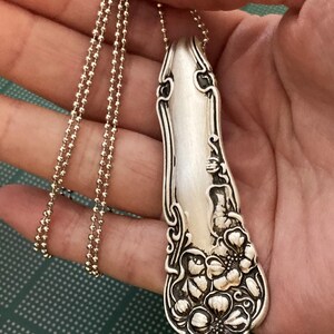 May include: A silver necklace with a pendant made from a vintage spoon. The spoon has a floral design and is attached to a silver ball chain.